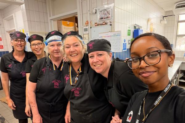 A cheerful group selfie of six kitchen staff in a tiled commercial kitchen. Wearing black uniforms, aprons and caps or hairnets, they stand close together smiling for the camera, conveying teamwork and camaraderie.