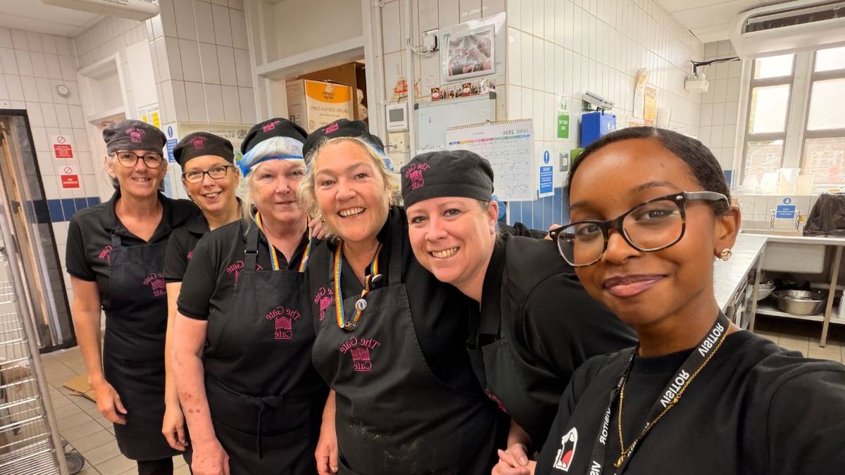 A cheerful group selfie of six kitchen staff in a tiled commercial kitchen. Wearing black uniforms, aprons and caps or hairnets, they stand close together smiling for the camera, conveying teamwork and camaraderie.