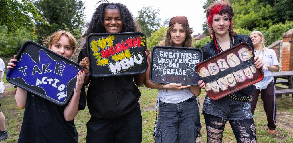 Four young people stand outdoors holding decorated serving trays painted with bold slogans — including "Take Action" and "Greatness Wasn't Raised on Grease" — smiling at the camera. They wear casual, eclectic clothes at a creative, activist-style gathering in a park.