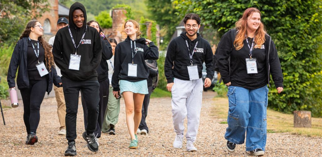 A group of young adults walking along a gravel path outdoors, smiling and chatting. They wear black hoodies, lanyards with badges and casual pants or shorts. Greenery and a brick building appear in the background.