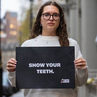 A young woman with long curly hair is holding a sign that reads 'show your teeth'