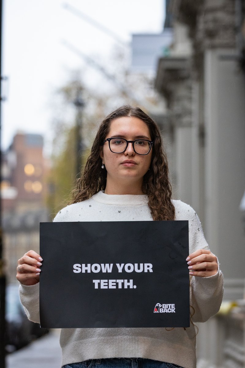 A young woman with long curly hair is holding a sign that reads 'show your teeth'