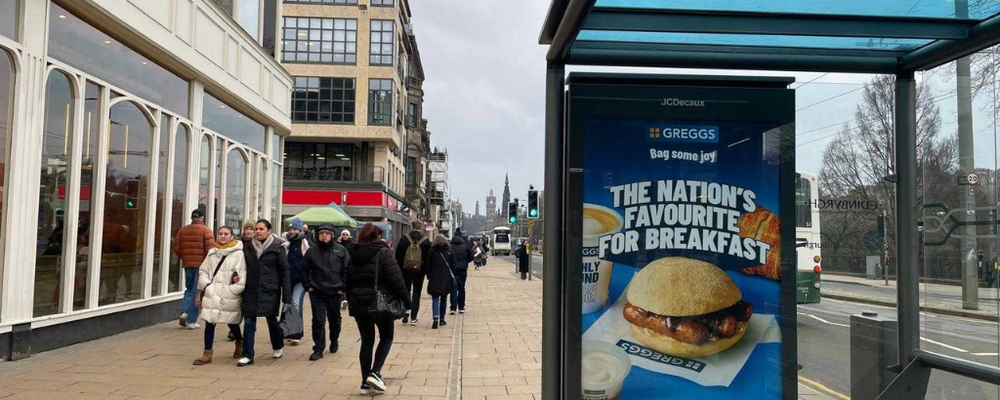 A busy street in Edinburgh featuring a bus stop that has a Greggs ad on the side billboard.