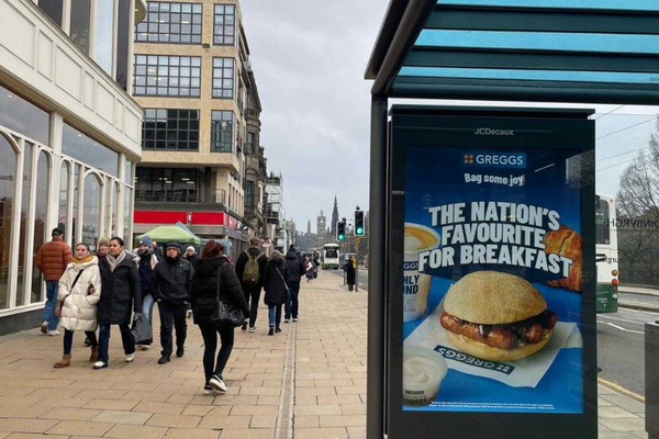 A busy street in Edinburgh featuring a bus stop that has a Greggs ad on the side billboard.