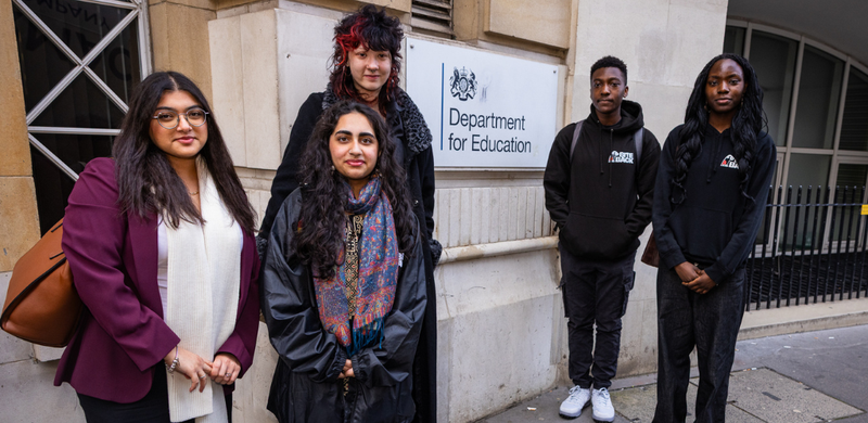 Five young people standing together in front of a Department for Education sign, wearing coats, hoodies and scarves, posing for a group photo.