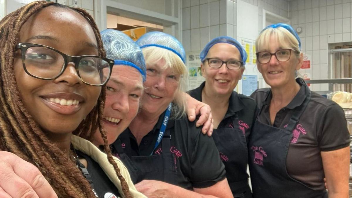 A selfie of five smiling women in a commercial kitchen — a woman with glasses and braided hair in front, and four staff behind wearing black polos, aprons and blue hairnets, with tiled walls and food trays in the background.