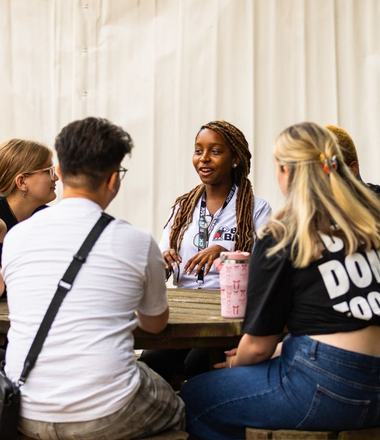 A diverse group of young people sit around a round wooden picnic table outdoors under a tent. A woman with braided hair speaks animatedly while the others listen attentively; several wear black shirts with bold white text.