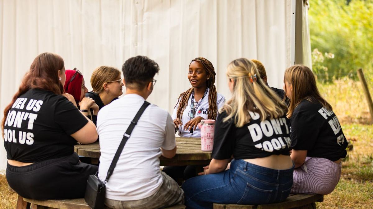 A diverse group of young people sit around a round wooden picnic table outdoors under a tent. A woman with braided hair speaks animatedly while the others listen attentively; several wear black shirts with bold white text.