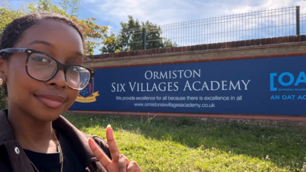 A person wearing glasses smiles and flashes a peace sign in a selfie taken outside on a sunny day, standing in front of a blue sign that reads "Ormiston Six Villages Academy" with grass and a fence visible behind.