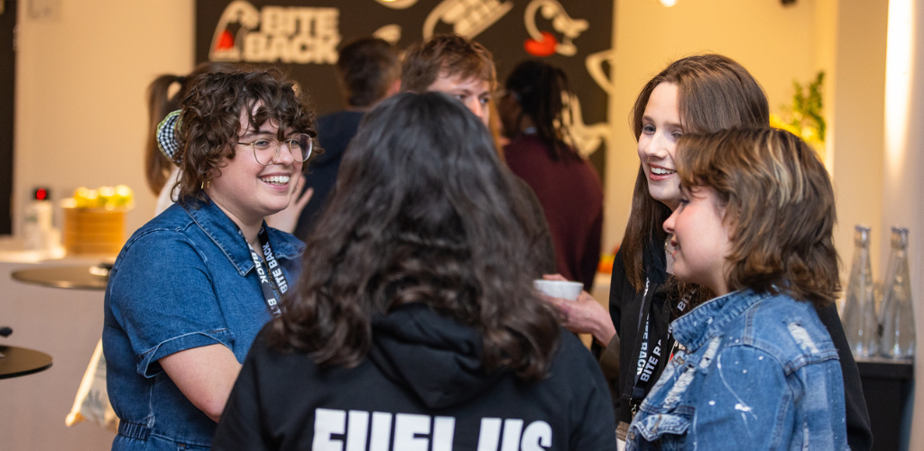 A Bite Back staff member is talking to three of Bite Back's young campaigners at a Bite Back event, they are stood around a table smiling