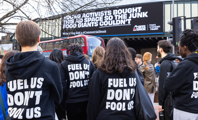 A group of Bite Back activists are looking up at a huge billboard in London that reads young activists bought this ad space so that junk food giants couldn't. They have their backs to the camera and their hoodies read fuel us don't fool us