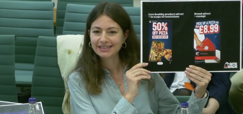 A photo of Bite Back's Head of Policy and Research Nika during the evidence session. She is smiling and holding up a printed graphic which shows an example of the loopholes in the upcoming junk food advertising legislation