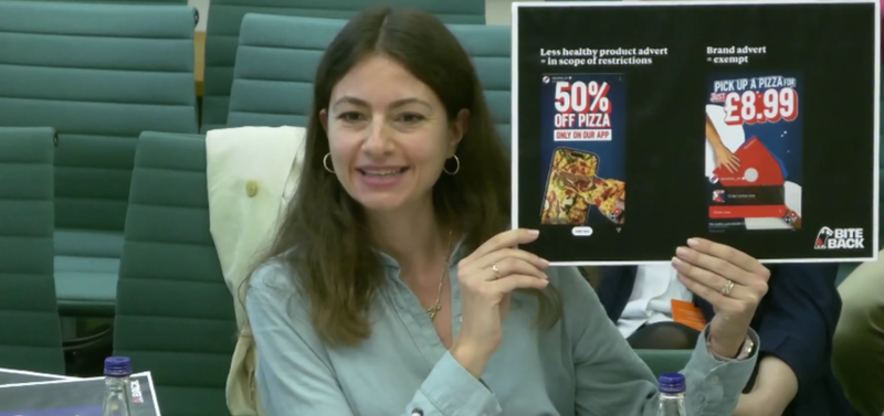 A photo of Bite Back staff member Nika during the evidence session in the House of Commons. She is smiling and holding up a printed graphic which shows an example of a loophole in the upcoming junk food advertising legislation