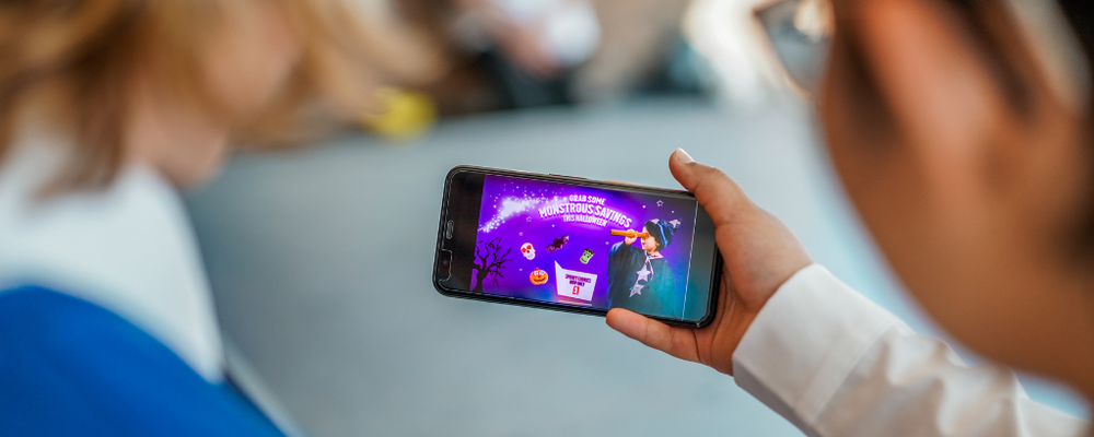 A photo taken over the shoulders of two children, one of them is holding a smartphone that features a Halloween-themed junk food advert, it reads grab some monstrous savings this halloween