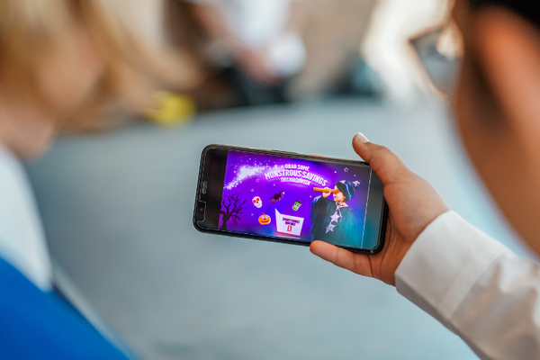 A photo taken over the shoulders of two children, one of them is holding a smartphone that features a Halloween-themed junk food advert, it reads grab some monstrous savings this halloween