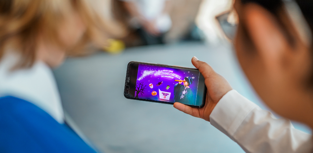 A photo taken over the shoulders of two children, one of them is holding a smartphone that features a Halloween-themed junk food advert, it reads grab some monstrous savings this halloween