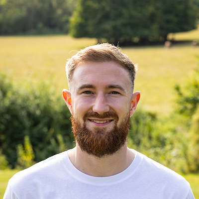 Andy a young white man wearing a white shirt with blonde hair, a kind smile and a bushy beard is standing in front of a sunsoaked field.