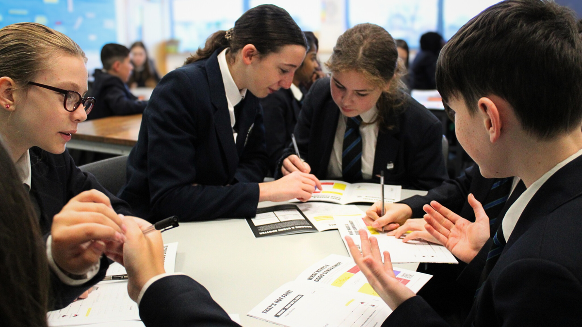 A group of students in school blazers and ties sit around a table in a classroom, leaning over worksheets and pens, focused on discussing and writing notes together.