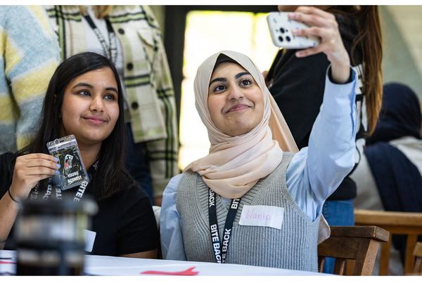 Two young women sit at a table smiling as they take a selfie — one wearing a light hijab holds up a smartphone, the other holds a lanyard card. They look happy and engaged in a busy indoor setting.