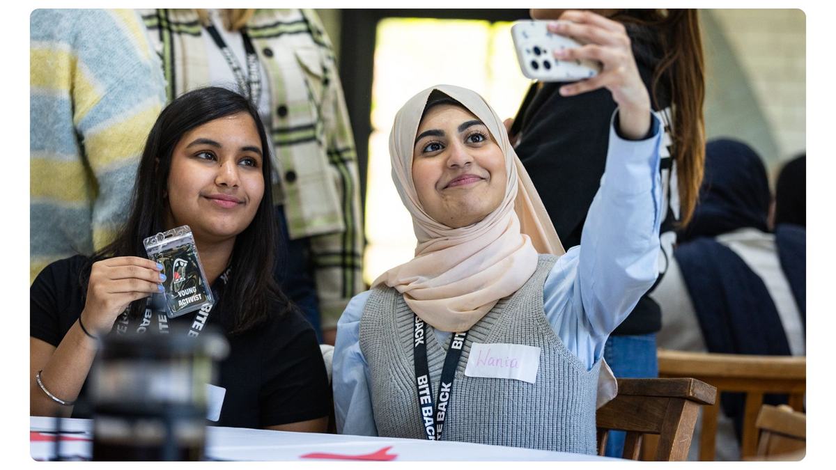 Two young women sit at a table smiling as they take a selfie — one wearing a light hijab holds up a smartphone, the other holds a lanyard card. They look happy and engaged in a busy indoor setting.