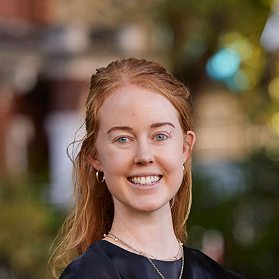 Caitlin, a young white woman with bright red hair, vibrant green eyes and a lovely smile is standing in front of a blurred background of trees and houses. She is wearing a plain black shirt, two necklaces and hoop earrings.