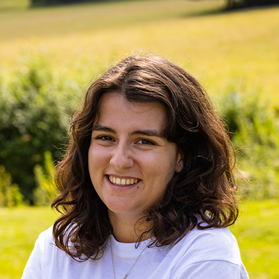 Carly is a young woman with an olive complexion. She has wavy thick brown hair, thick eyebrows and a lovely smile. She's standing in front of green and yellow fields wearing a plain white shirt.