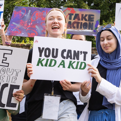 Bite Back activist Clemmie is smiling and holding a protest sign that reads You Can't Kid A Kid, she is surrounded by other signs and activists