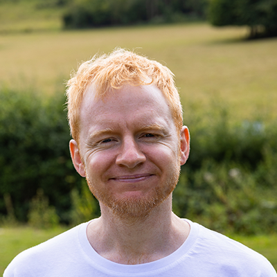 Dan a white man with redblonde short hair, freckles, blue eyes and a lovely smile stands in front of a scenery of fields and greenery.