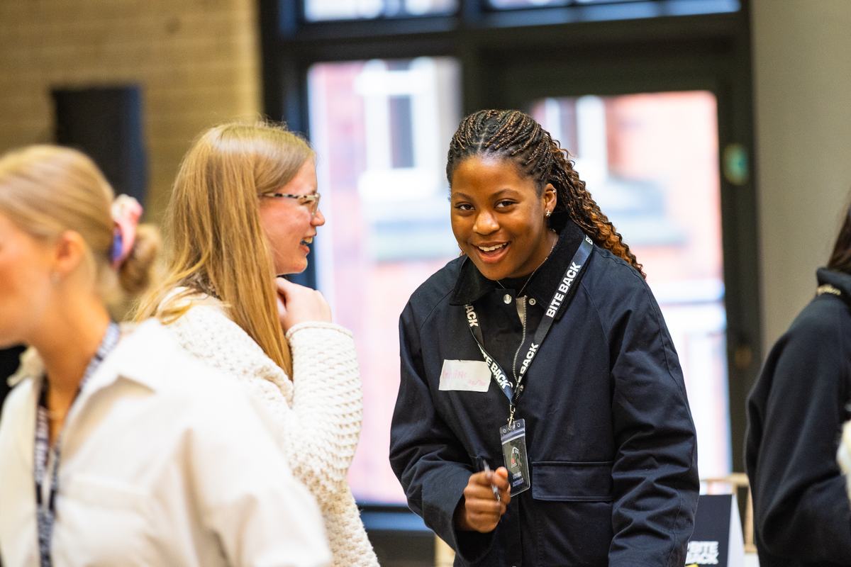 A candid indoor scene of young people chatting and laughing. The focus is a smiling Black woman with braided hair wearing a dark jacket and a "BITE BACK" lanyard, talking with a blonde woman in a cream sweater. Background is softly blurred.