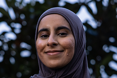 Faheemah is a young petite brown woman wearing a greyish brown hijab slung across her shoulders.