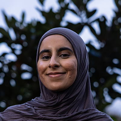 Faheemah is a young petite brown woman wearing a greyish brown hijab slung across her shoulders.