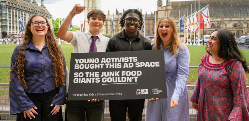 Five Bite Back activists stand smiling in front of Parliament in London, they are holding a sign represeting Bite Back's banned billboard which reads Young Activists Bought This Ad Space So The Junk Food Giants Couldn't