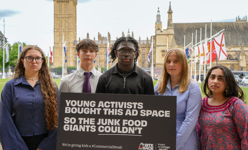 Five Bite Back youth activists stand in front of the Houses of Parliament with serious expressions. They are holding a copy of Bite Back's banned billboard which reads young activists bought this ad space so the junk food giants couldn't