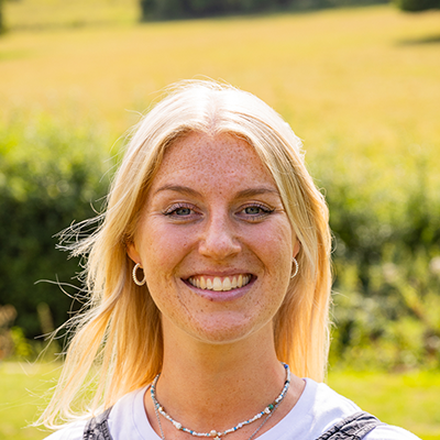 Hannah is a young white woman with a big smile, pale blond hair, freckles across her cheeks and vibrant blue eyes. She is wearing a white shirt and standing in front of sunsoaked fields.