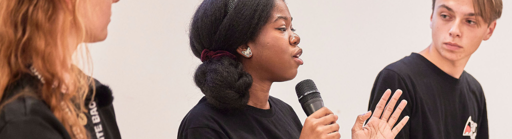 Header Bite Back activist Derrin is talking on a panel at a Bite Back campaign launch event. She is speaking confidently into a microphone, two other panel members are looking at her and listening.