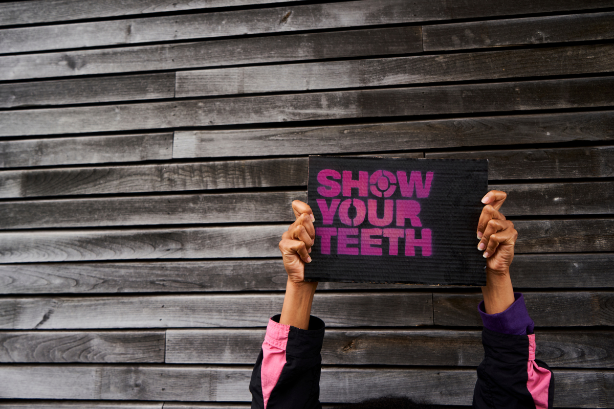 Two brown hands holding up a protest sign on which is spray painted 'Show Your Teeth' in purple on black background.