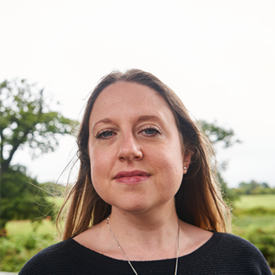 Gina a staff member with Bite Back. She is a white woman white a round face and long blonde hair. She is wearing a black jumper with a silver pendant necklace. She's standing in front of a cloudy sky and trees and is wearing a serene expression on her face.