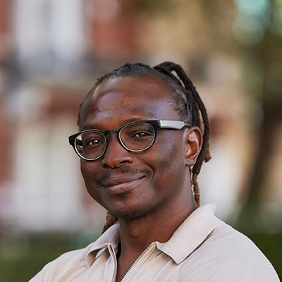 Henry a black man wearing a white shirt with a big lovely smile and his dreads pulled back into a pony tail is standing in front of green and yellow fields.