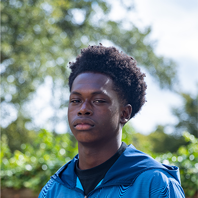 A young black man with curly hair styled in a fade is looking into the camera with a serious gaze.