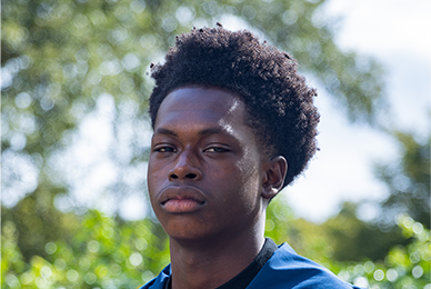 A young black man with curly hair styled in a fade is looking into the camera with a serious gaze.