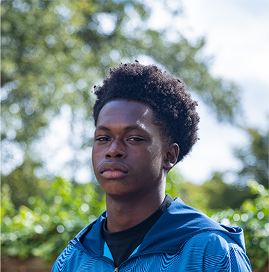 A young black man with curly hair styled in a fade is looking into the camera with a serious gaze.