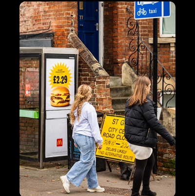 A photo of two young people walking past an ad for a junk food product