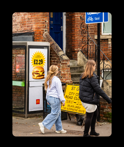 A photo of two young people walking past an ad for a junk food product