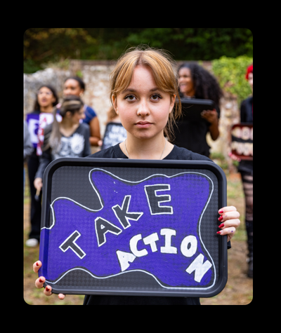 A young blonde woman is standing in a field holding a painted school dinner tray that reads 'take action'