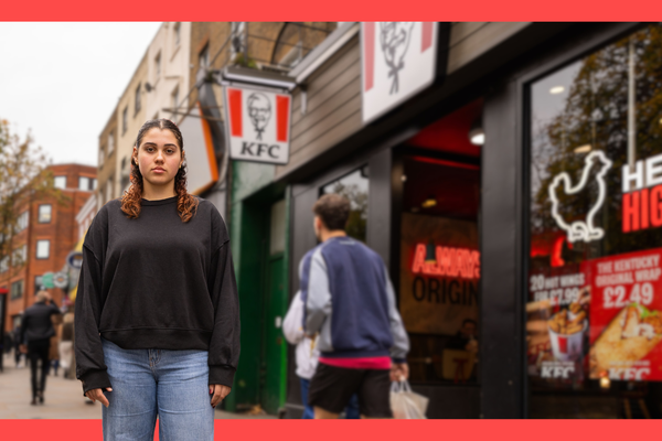 A young woman stands on a busy city sidewalk in front of a KFC restaurant. She wears a black sweatshirt and jeans, looking at the camera while pedestrians pass by and the KFC sign and posters appear behind her.