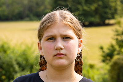 A young white woman with blonde hair in two braids is looking into the camera with a serious gaze.