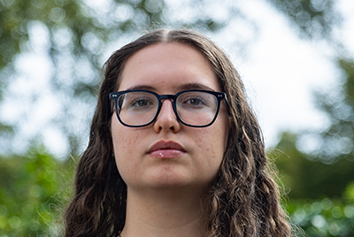 A young white woman with curly brown hair and glasses is staring sternly into the camera.