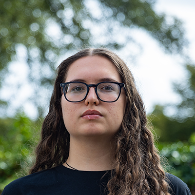 A young white woman with curly brown hair and glasses is staring sternly into the camera.
