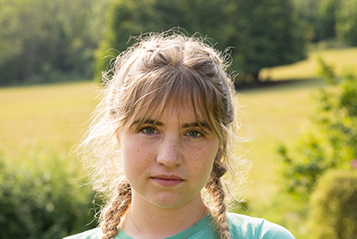 A young white woman with blonde hair pulled into braids is looking at the camera with a serious expression.