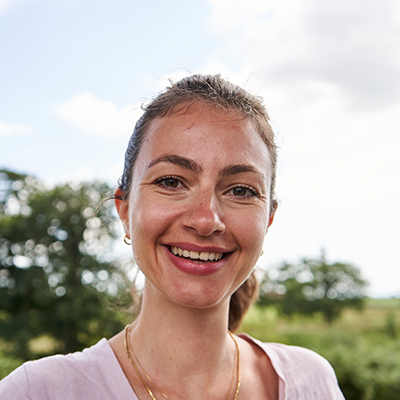 Nika is a young white woman with long brown hair tied back into a ponytail and small hoop earrings. She is standing in front of a scenery of beautiful blue sky and trees and wears a pale pink shirt with a golden necklace.